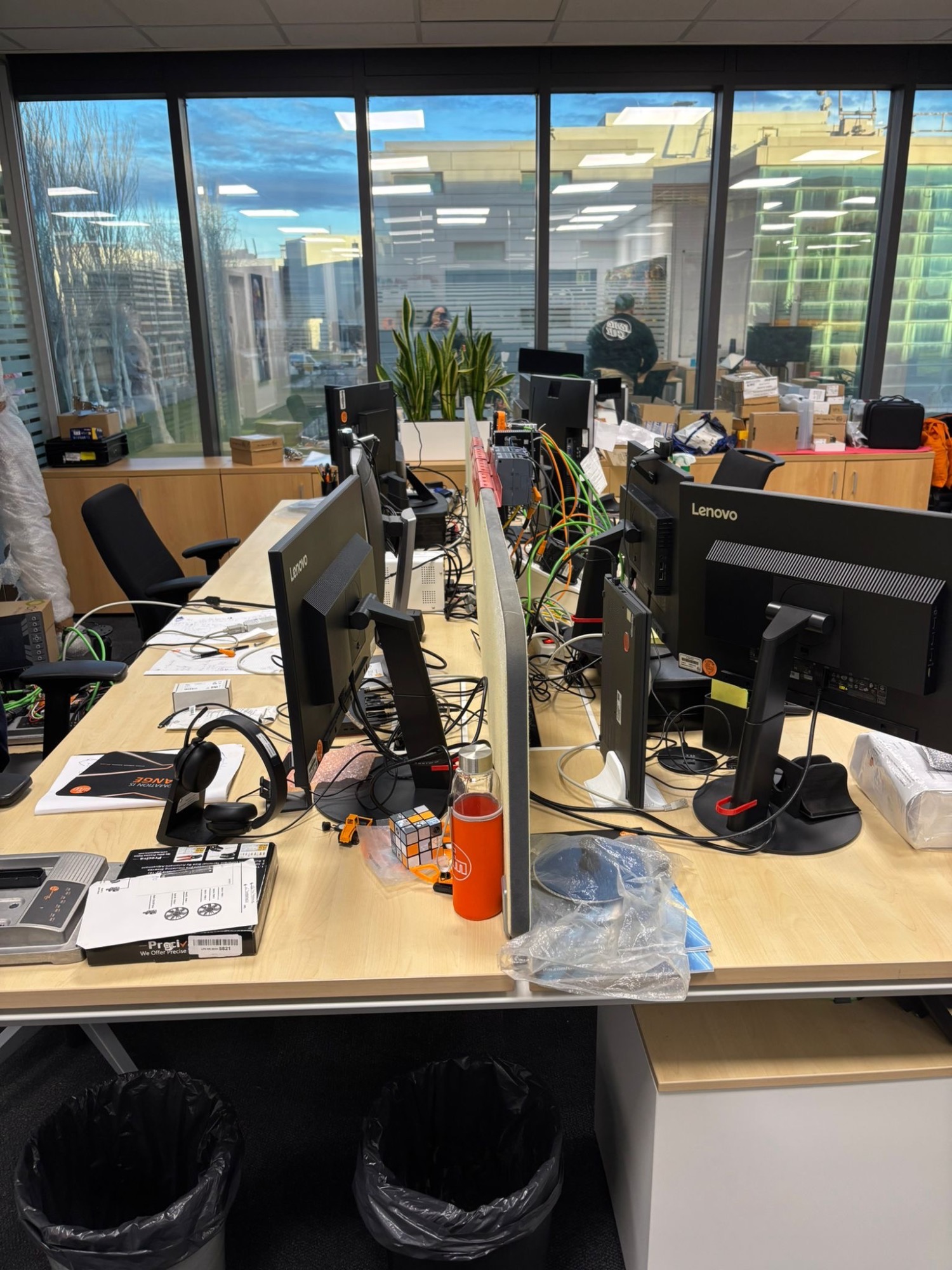 Escritorios de oficina con monitores, papelería y plantas, vista de un entorno laboral moderno.